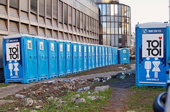 Eine (ordentliche - mehr als 15 Stück) Reihe von blauen toitoi Toilettenhäuschen. Auf diesem Gelände liegen aber sehr viele leere Plastik-Getränkeflaschen (Müll) am Boden. 