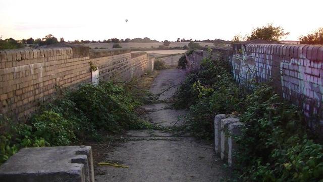 view along a narrow disused Victorian road bridge over a railway not long before sunset. some graffiti on the right hand wall. a couple of concrete blocks for some reason. there is no road leading to it any more. maybe to stop farm vehicles, maybe it wouldn't handle the weight.  over grown with brambles. the blackberries tasted bad also.