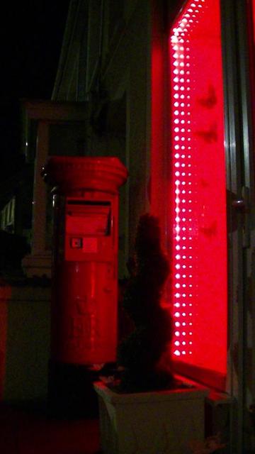 a red british EiiR red post box next to a shop window at night, but also illuminated red from red LED lights around the inside of the shop window. window has  butterflies on the inside wall.