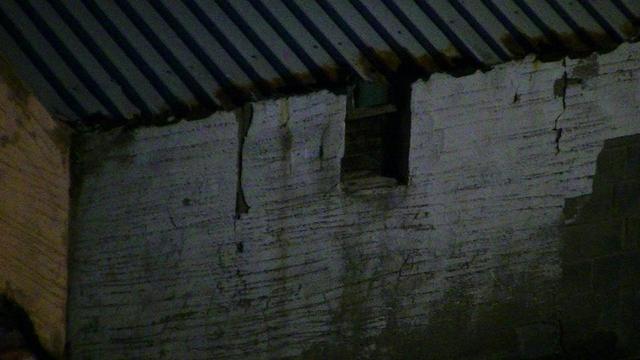 looking down at a lime rendered breeze block building with a tiny dark window at nigh time. it has grey steel box profile roofing that has rusted away at the ends. some of the rendering has cracked an or crumbled away. 