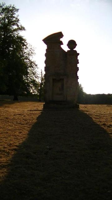 remains of a gate post in the middle of a field. one of a pair. the sun is behind it casting a long shadow towards the camera. the gate post is built of limestone blocks to a height of about 5 to 6 meters. it has a bit of a slant on a pier capstone on the left, and a limestone ball final on the right of this large single post. it has an alcove to sit in at the bottom where i found some antique graffiti dated 1878. the road no longer exists. 
it is just a field of brown grass.And the snap was fr…