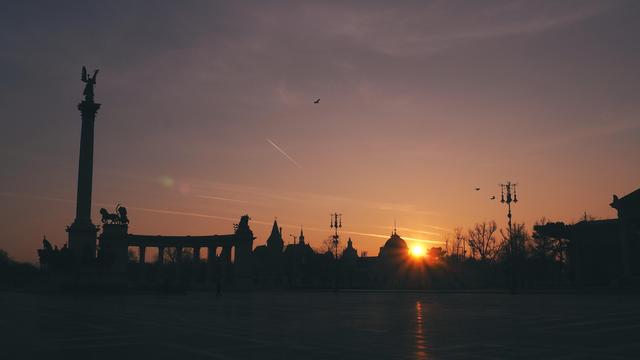 Ein großer freier Platz mit einer Säule links auf der ein Engel die Spitze bildet, daneben einige Dächer und Leuchten, alles als Scherenschnitte abgebildet, rechts unten die Sonne bei Sonnenaufgang am Horizont. Am rötlichen Himmel dünne Schleierwolken. 