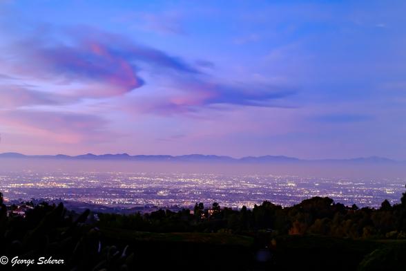 View of the lights in the LA basin, with a layer of fog, and the mountains in the background.  Sunset has lit up the clouds in colors of dark blue and pink.