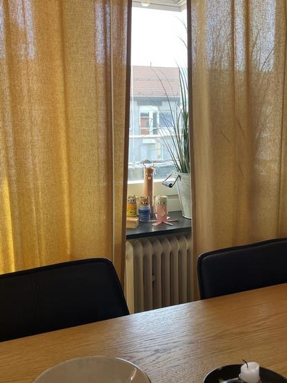 A cozy interior view featuring yellow curtains, a wooden table with a plate, and a black chair. In the background, a window shows a verdant plant, a whimsical figure, and colorful decorative items on a windowsill, while a radiator is visible