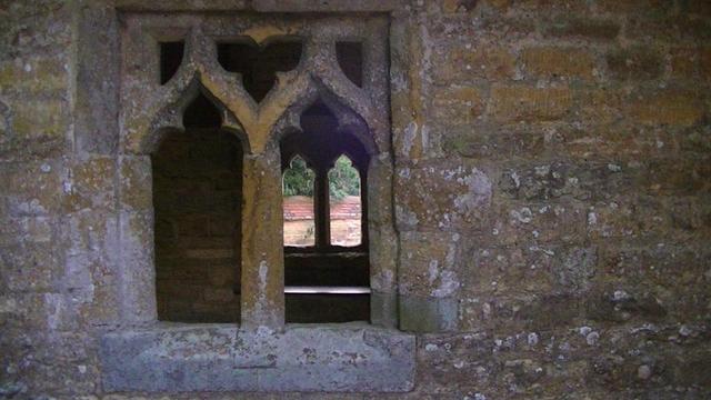looking west through a limestone church porch 2 arch window with no glass, and through the window on opposite side of the porch, also no glass. there is a limestone wall topped with red terracotta tiles under a tree through that far window. this is an old church with parts ranging from 13 to the 17 century. same church as the gargoyle i took a snap of yesterday. the walls are a dark limestone, covered in lots of white lichen.