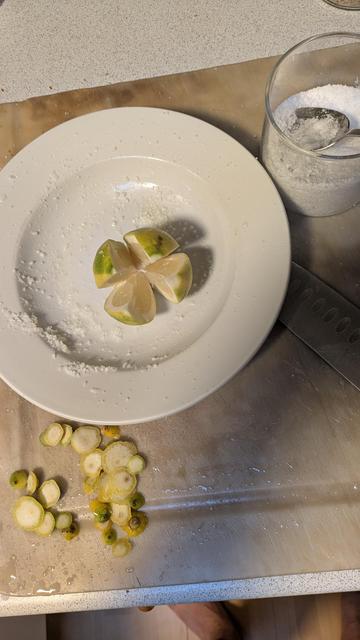 Foto af en hvid tallerken med en enkelt citron, som er skåret op næsten helt i bund, men de fire spalter hænger stadig sammen. Citronen bliver som det næste fyldt med en spiseske groft salt. Lidt salt kan også ses rundt omkring i tallerkenen,