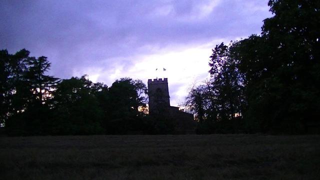view across a field towards a silhouetted church that the sun sets behind. many different species of trees silhouetted either side. purple sky, with few white and orange bits. same church as yesterday. or maybe the day before now. anyway, it is a church with a tower.