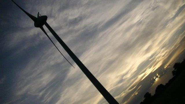 maybe an hour later, sun is setting, the snap is at an angle to get some of the horizon in and the turbine. it's a silhouette of the wind turbine in the foreground. no room to walk backwards any further. orangey brown sky, clouds obscuring the sunset. closer clouds are wispier.