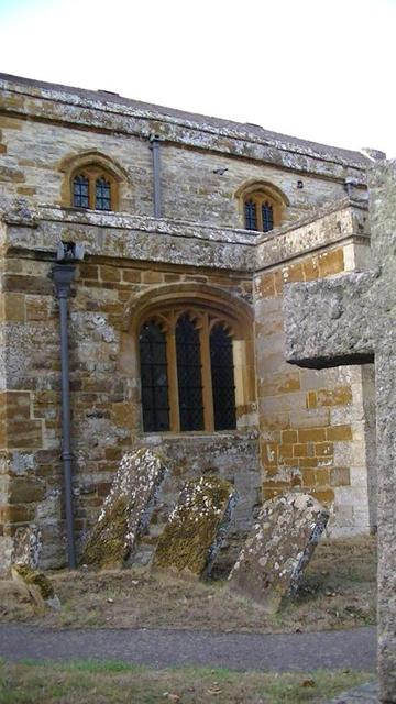 south wall of a limestone and ironstone medieval church with leaning lichen covered limestone grave stones. above the grave stones is a mouth like isle widow, then above is two eye like clerestory windows. another snap from yesterday.