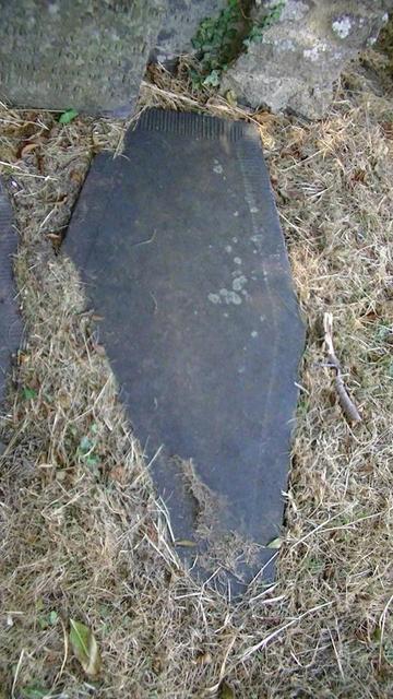 looking down at a heavy coffin shaped slate slab on top of a grave in front of a headstone. it has carved borders of like radiating lines. it is one of a pair of graves. it is surrounded by brown grass.