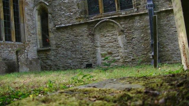 a door that has been blocked off, maybe centuries ago, on the south wall off a limestone medieval church to the right of the south transept, of which part of the window is in view. there is a tall thin arch window to the left of the door and the bottom half of a window above. a few grave stones side on. the grass is starting to grow green again after this hot, dry summer turned it brown. view from top of chest tomb. 