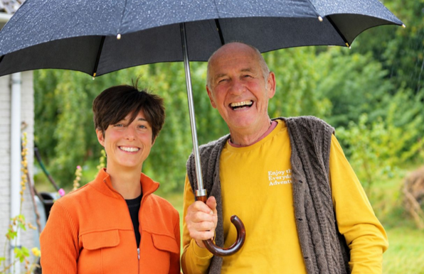Foto af Tatiana og Peter. De står under en sort paraply, da det er udenfor og det regner.
Hun er iført en orange frakke, han har en gul t-shirt med en uldvest på. Begge smiler bredt.
Foto by Katrine Hedegaard