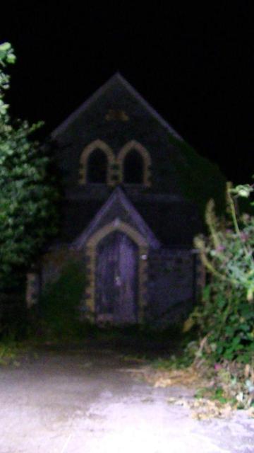 in the dead of night. in torch light. a narrow house. the upper floor has 2 arched windows close together, well.. next to each other, touching. (looks like eyes), and the point of a steeply pitched gable roofed porch, lines up between the windows, it has a gothic arch door, looks like a dogs nose. it is very noisy and blurry snap, it's shit but i prefer this one. to the focused not so noisy one that i also got. also btw it is viewed from the entrance of a narrow drive lined with small trees tha…