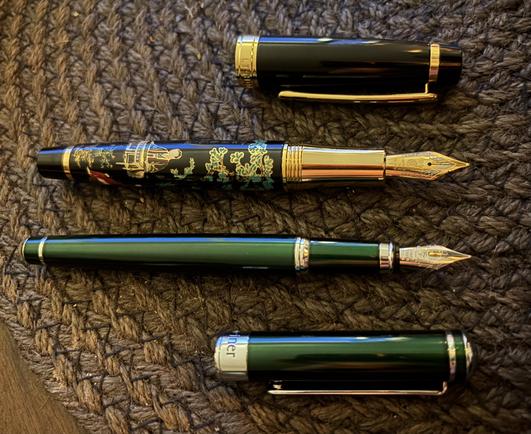 Two fountain pens on a table. One, a jade green design and a silvery nib - and the other black with a traditional Chinese painting on it in green, gold and red and a golden nib. 