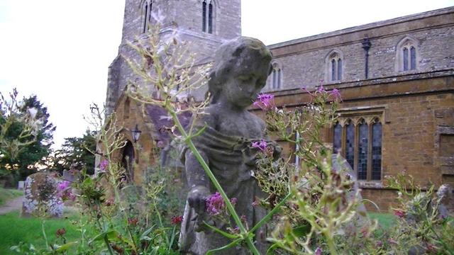 a stone statue of a little girl, marking a grave, looking down, amongst red valerian flowers. church in the background from the south. limestone naive and tower. ironstone aisle walls. 13th and 15th century apparently. sky covered in white cloud.