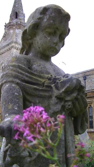 close up of the stone statue of a little girl, marking a grave, looking down, amongst red valerian flowers. limestone church steeple in background. very fine cobweb draped from her cheeks. she looks very sad.