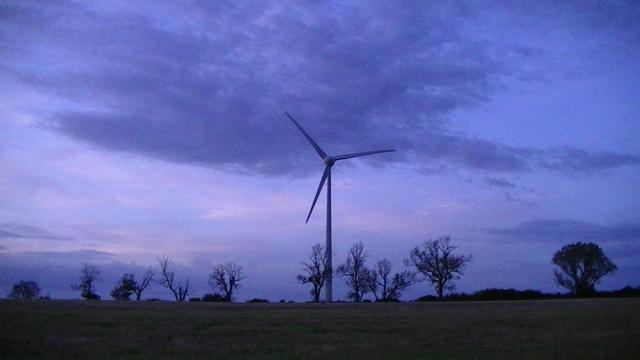 looking across a field at dusk toward a wind turbine. there is a row of trees, most of them have no leaves, prolly dead then, as i only did the snap a couple of weeks ago. or maybe less. the sky is a very purpley blue. the field is got a purple vibe too. trees are like a silhouette. also a few clouds of similar shades to the sky.