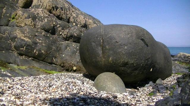 a big smoothed rock on a rocky sea shore. there is a bit of sea in the distance on the right. the rock has some discolouration that looks like an eye, vertical line of quartz in the middle of its face. looks like some sort of stone plushy. shell fragment shale or whatever in the foreground. it was in one of the heat waves last month, you know, clear skies strong shadows. and there are barnacles and green stuff on the rocks in the background, well its one giant smoothed outcrop.