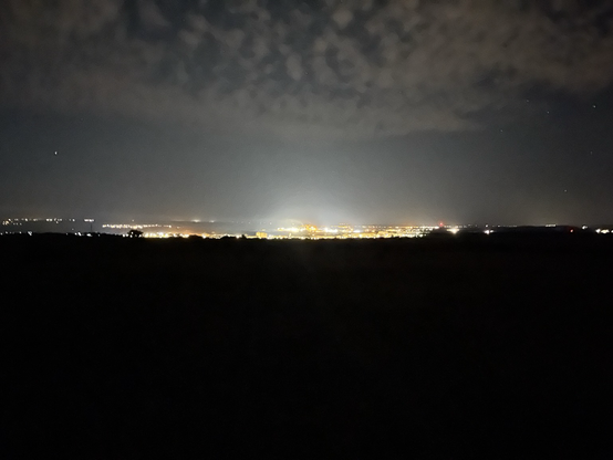 Distant industrial city view; black foreground, lights above, night sky with clouds above that 