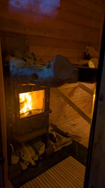 Foto af en brændende saunaovn med lystigt flagrende flammer, taget igennem glasdøren.
Der ligger stykker af brænde under ovnen.