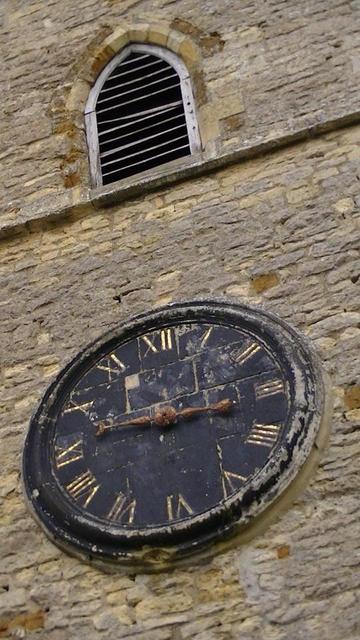 a arch bell louvre church window above a not working black paint flaking clock gold roman numerals, rusty hands. limestone rubble church.