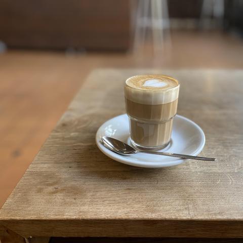 A steaming cup of cortado on a wooden café table in a Berlin café. A darker wooden floor is visible behind the table and a brown sofa and chairs are out of focus in the background.