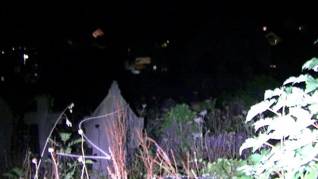 some graves at close to midnight in an overgrown graveyard on the side of a hill in torchlight. and the lights of a town is in the background lower down the hill. the snap is a bit noisy and blurry even by my low standards, lol.