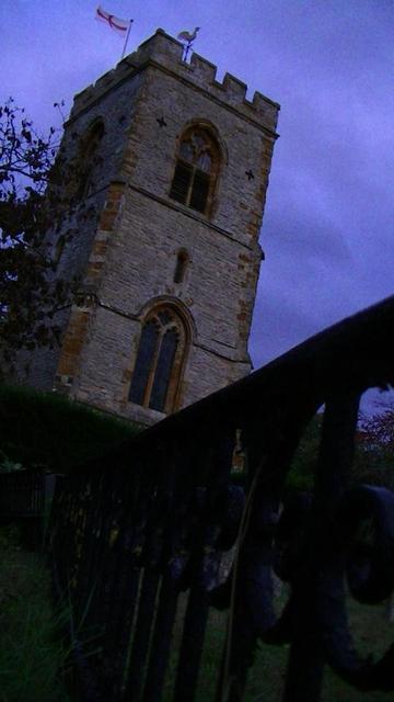 looking along cast iron railings towards a medieval church tower. railings with like scrolls. it is a limestone rubble tower with odd bits of ironstone on the corners. big top arch window with wooden slatted quatrefoil. wooden bell louvres. bottom window same as top only with leaded glass. small narrow window in the middle. weather vein, flag etc. purple sky just after sunset.