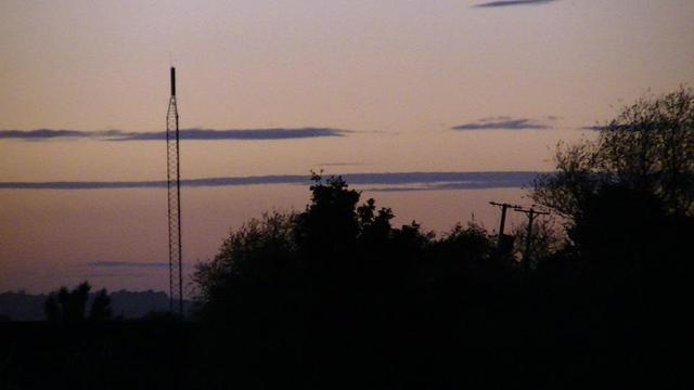 zoomed into the transmitter sticking out above trees. 2 wooden electricity poles leaning badly to the right.