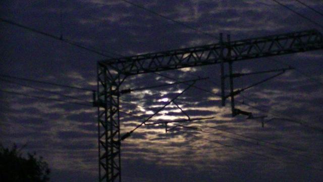 blurry moonlight shinning through small gappy clouds, at night, behind overhead electrified railway cables and their mast thingy for hanging them.