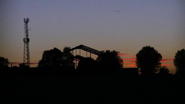 dusk sky with orange red clouds reflecting the sun from below the horizon, behind the silhouette a transmitter, trees and the skeletal steel frame of a building.
