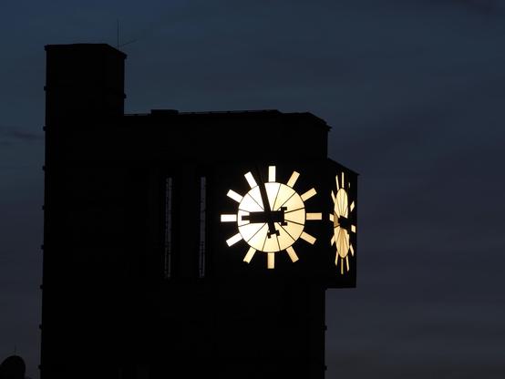 Das obere Ende eines turmartigen Gebäudes mit einer beleuchteten Uhr. Der Himmel ist sehr dunkel.