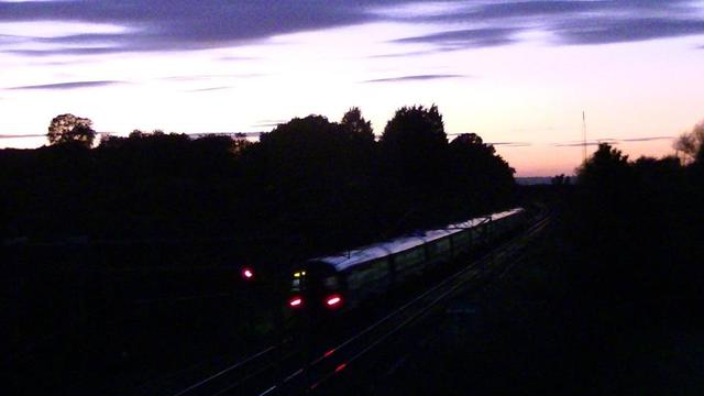 blurry snap of a train at dusk. deep blue clouds. glowing electric blue sky fading to orange on the tree lined horizon. the snap is from the right side of a bridge. so the train track starts from the left of the frame then the train is dissapearing into the distance on the right. red lights at the back. actually the signal looks red too.... that can't be good. well anyway them red lights are reflected in the rails, because it's dark down there.
