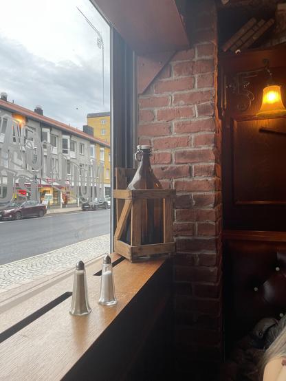 A cozy restaurant corner featuring a brick wall and wooden window sill. There is a brown wooden crate and a glass bottle on the sill, alongside two salt and pepper shakers. Outside the window, city buildings and a quiet street are visible under a cloudy