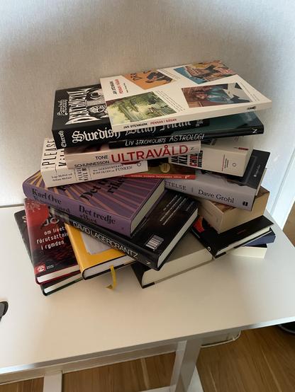 A disorganized stack of various books piled on a table. The titles and authors include a mix of fiction, non-fiction, and possibly biographies, with some written in different languages. The background features a simple, light-colored wall.