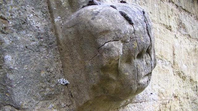 a crude and cracked carving of a human head at the bottom of a church window hoodmould. grey limestone. few bits of yellow and white lichen. dark moss growing out of the cracks at the top.