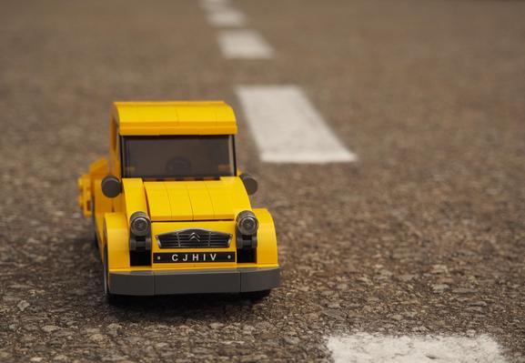 Ein gelbes Spielzeugauto (2CV) auf der Straße. Die weiße Leitlinie teilt das Bild. 