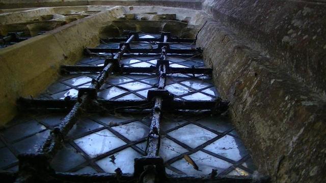 looking sharply up at a limestone arch of a church window with filthy leaded glass, cobwebs with some sycamore seeds stuck in them. posh square section iron support bars look blacksmith made. the horizontal ones have square protruding square eyes for the vertical bars. there was fleur de lis on the tops but not visible from this angle. the church prolly 14 century but this window is victorian.