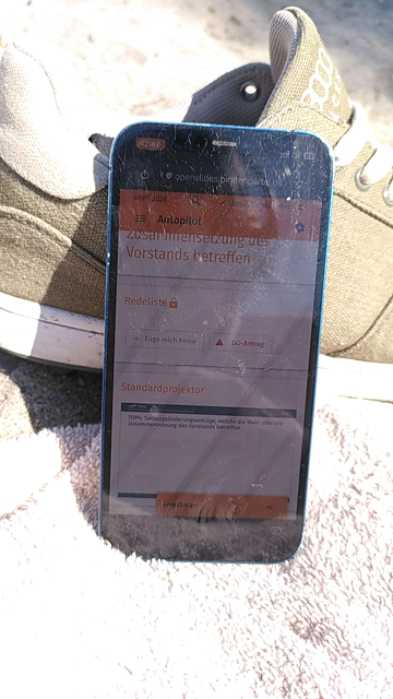 Ein Handy auf einem Handtuch am Strand, im Hintergrund Sonnenschirme und das Meer