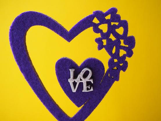 Zwei miteinander verbundene violette Herzen aus Moosgummi ausgestanzt, in der Mitte der Schriftzug LOVE auf gelbem einfärbigen Hintergrund