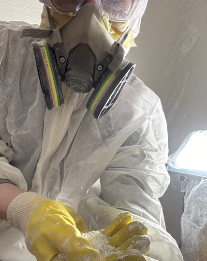 A person in a breathing mask with aerosol filters, protective glasses, clothing and yellow rubber gloves holding some white powder in their hand.
There is some plasting coverings over white walls and a LED torch in the background.