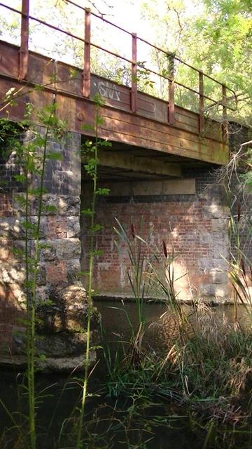 a bridge with red brick and stone piers over a river with bull rushes or what ever.
it has rusty 2 bar railings on rusty cast iron sides, on plate girders. not sure of the age. i am guessing early 20th century.
