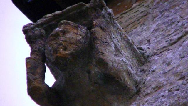 a stone gargoyle type carving beneath the guttering on the gable end of a medieval church. it has arms holding up something that is not there no more. maybe a sump or a spout to direct the rain water away from the wall. the weathered face looks very strained. must of been made of lead. prolly why it's not there no more.