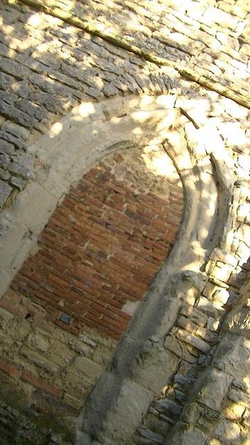 a pointed arch doorway at the bottom of a limestone rubble medieval church tower. the bottom third of the bricked up bit is made of large limestone blocks. some of the look red, like they were once part of a furnace or fireplace. then the top two thirds is made of red brick, looks like proper old Tudor red brick, or even older, they are not very deep in thickness. 