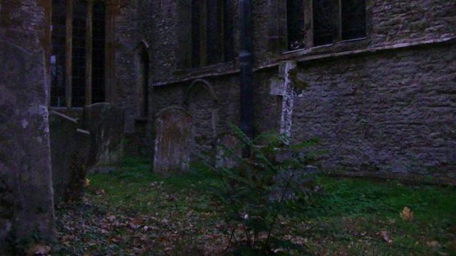 dusk at the corner of a small churchyard. some graves in foreground. lookin in the corner of the south wall, east wall of south isle. medieval limestone rubble church. bottom of leaded windows visible.