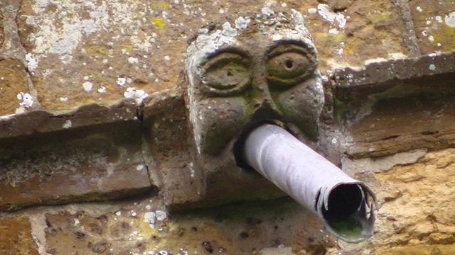 a grotesque face with lead pipe sticking out of its mouth. top of a limestone church tower. looks like crocodile teeth a the end of the pipe.