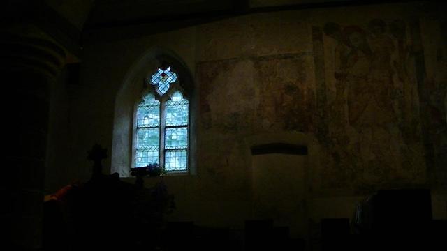 inside a dimly lit medieval church. two light arch leaded window with quatrefoil on the left, letting in dull light. st.Christopher mural on right. bricked up doorway in the middle 