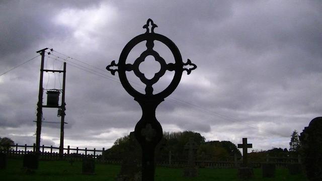 an iron headstone errm ironstone?! no. headiron, graveiron. well it's a gravestone size cast iron grave marker, in a graveyard surrounded by a wooden picket like fence. there is a couple of telegraph poles with a transformer high up in between in the background on the left with wires. some ivy on left pole, dark clouds in the sky.
