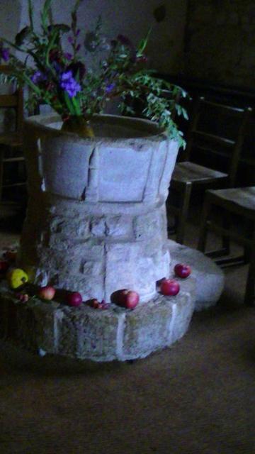 a medieval limestone font with flowers in the bowl. And fruit mostly apples around the plinth. some wooden chairs aside the font. it was a dimly lit corner of the church so i shined my torch but the picture is still noisy, tho i would not call the snap shite imo. looks like they still have harvest festival here. it was a big thing in rural UK when i was a kid. fucking long time ago now, lol