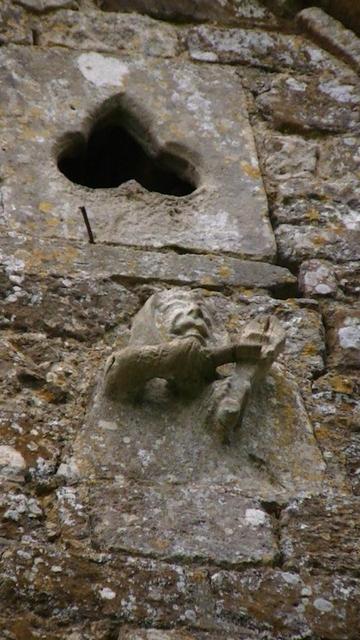 a limestone carving of what looks like someone playing a stringed instrument under a trefoil near the top of a medieval church gable end